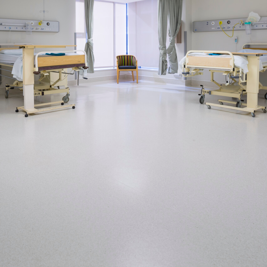 Quarto hospitalar com duas camas, cortinas, cadeira central e piso vin&iacute;lico claro.